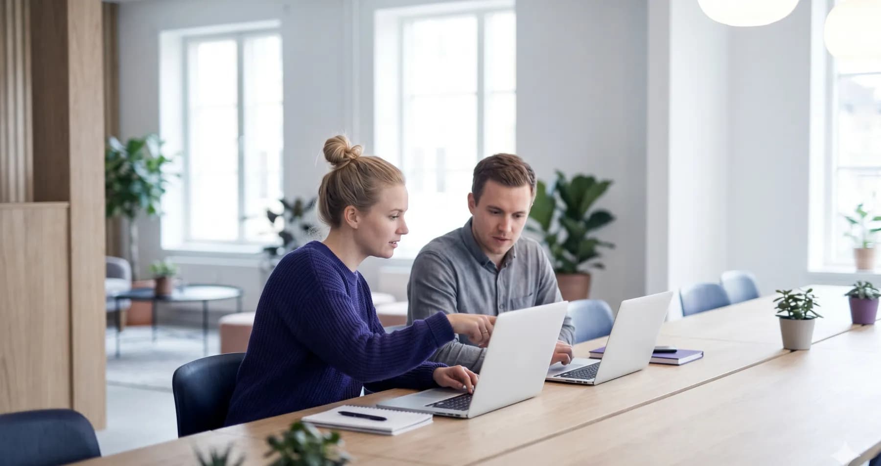 Två personer som samarbetar vid ett ljust träbord med laptops i en nordisk studio